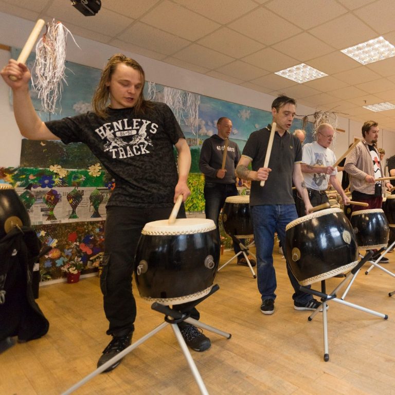 A group of men of all ages hitting large drums at a music wellbeing workshop